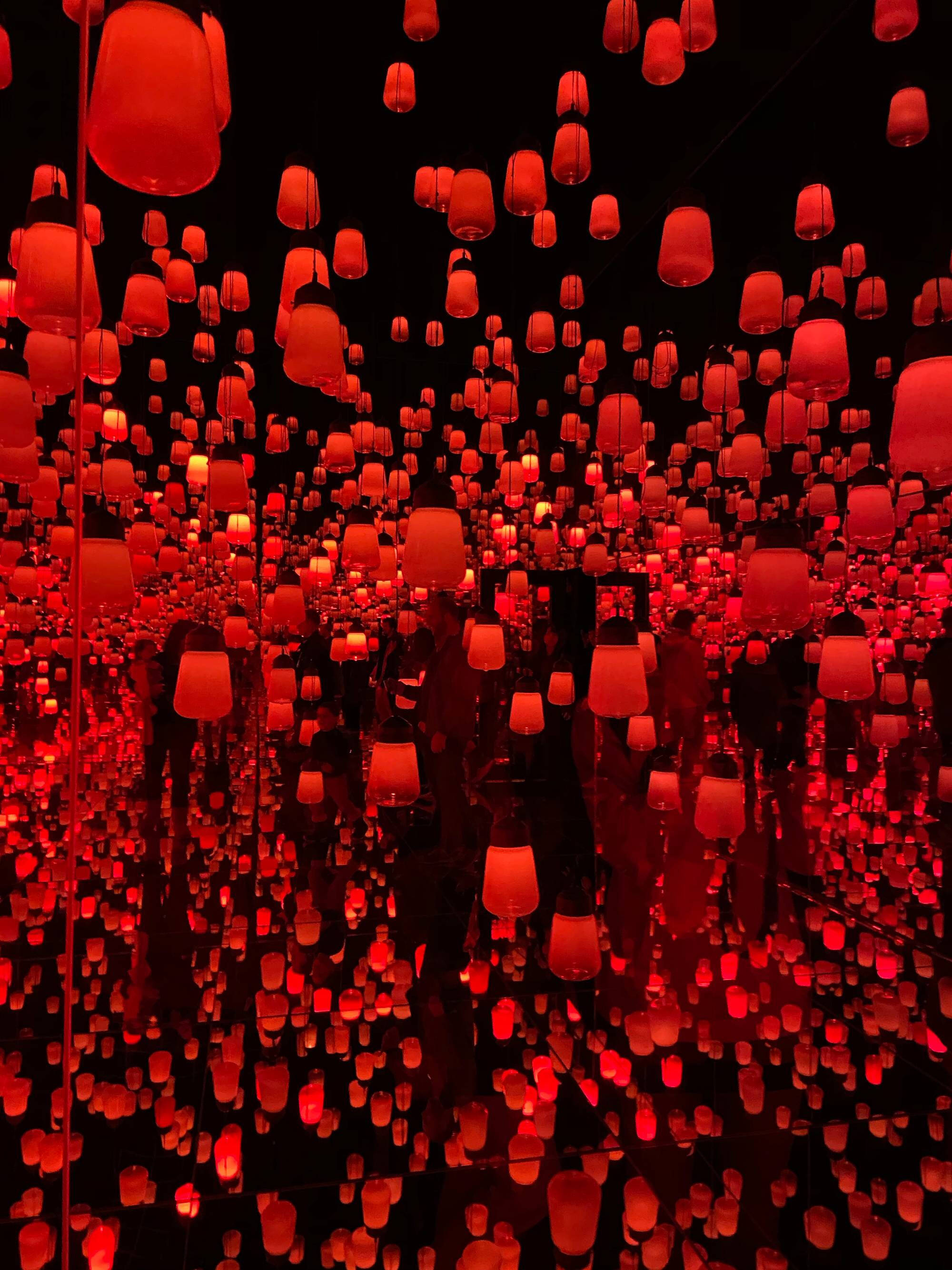 Colored photograph of red lights or lanterns in a mirrored space. Few people visible among the lights near the center of the image.